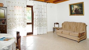 Un salon avec un plafond en bois, un canapé beige, une chaise en bois, une table recouverte d’une nappe en dentelle et un tableau accroché au mur.