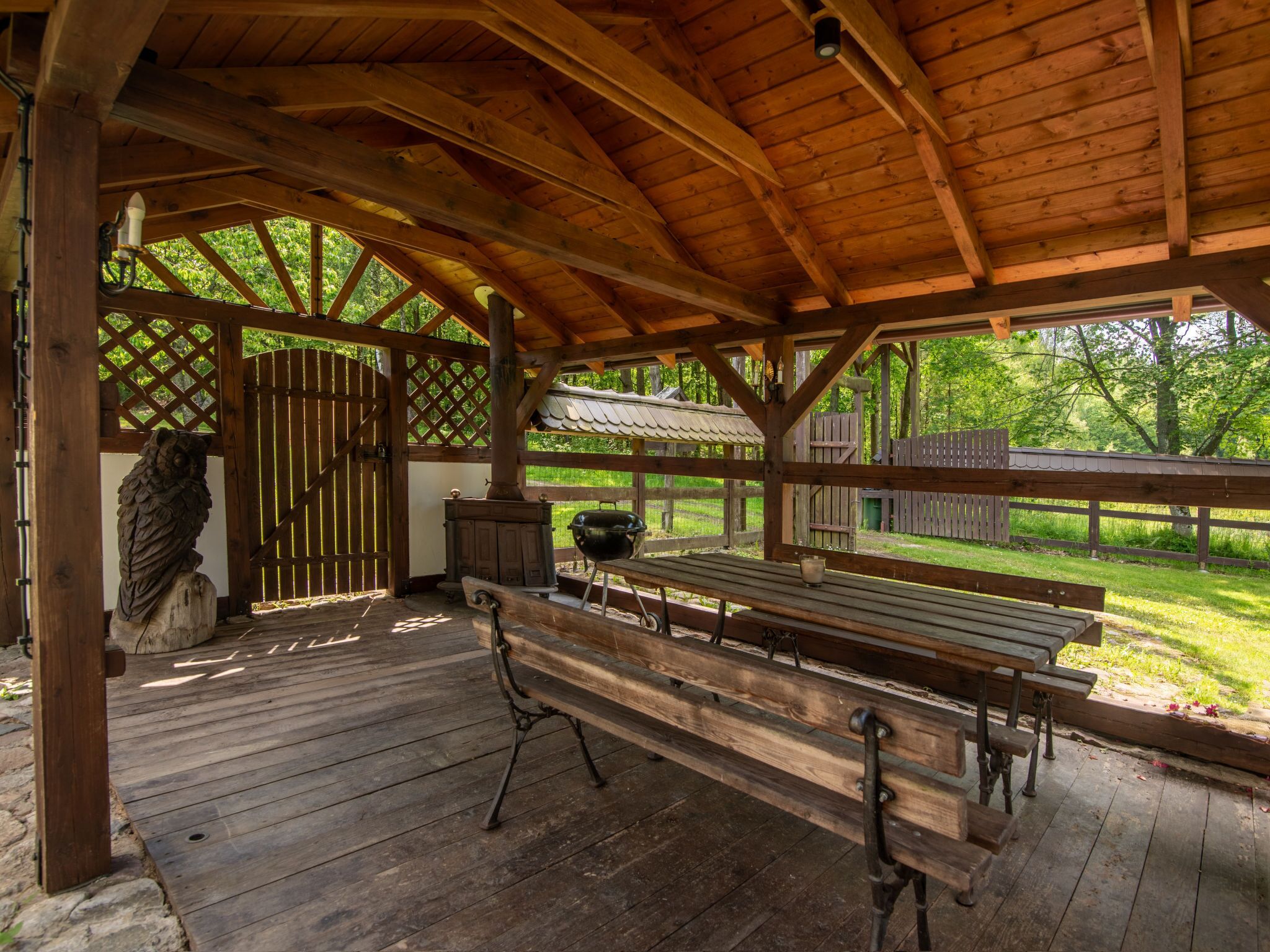 Un pavillon en bois doté de bancs et d’un espace barbecue, entouré d’une clôture en bois et de verdure.
