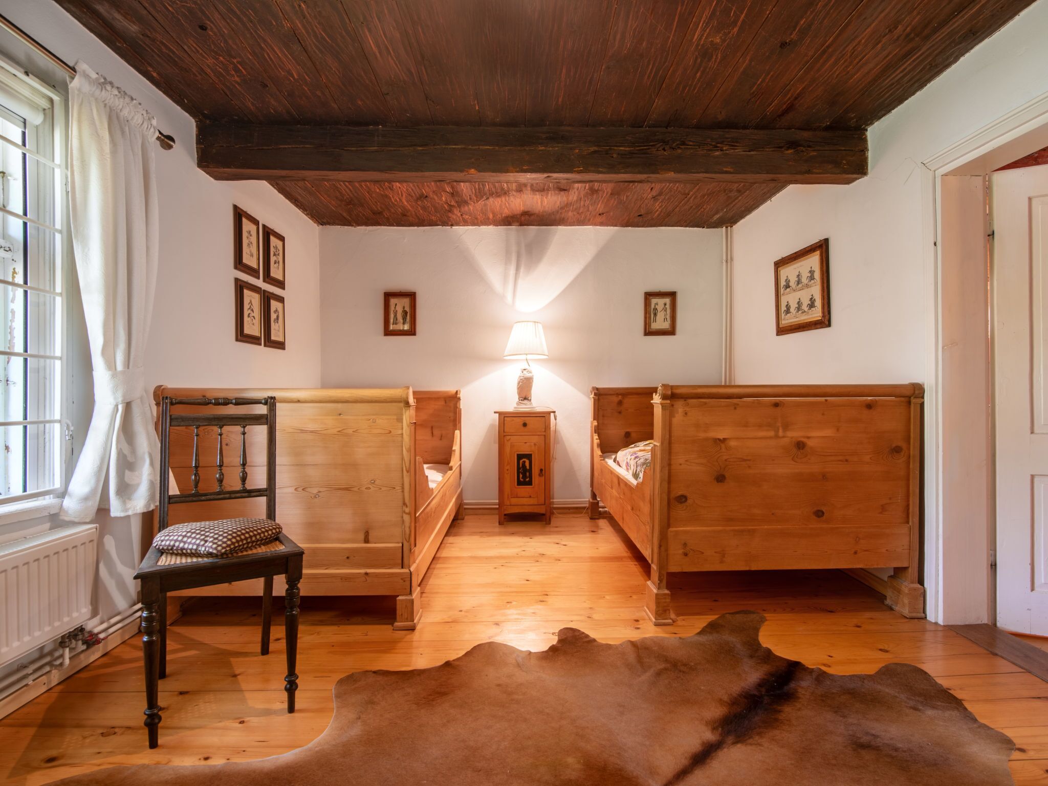 Una habitación con dos camas de madera, una silla, una lámpara y una alfombra de cuero de vaca.