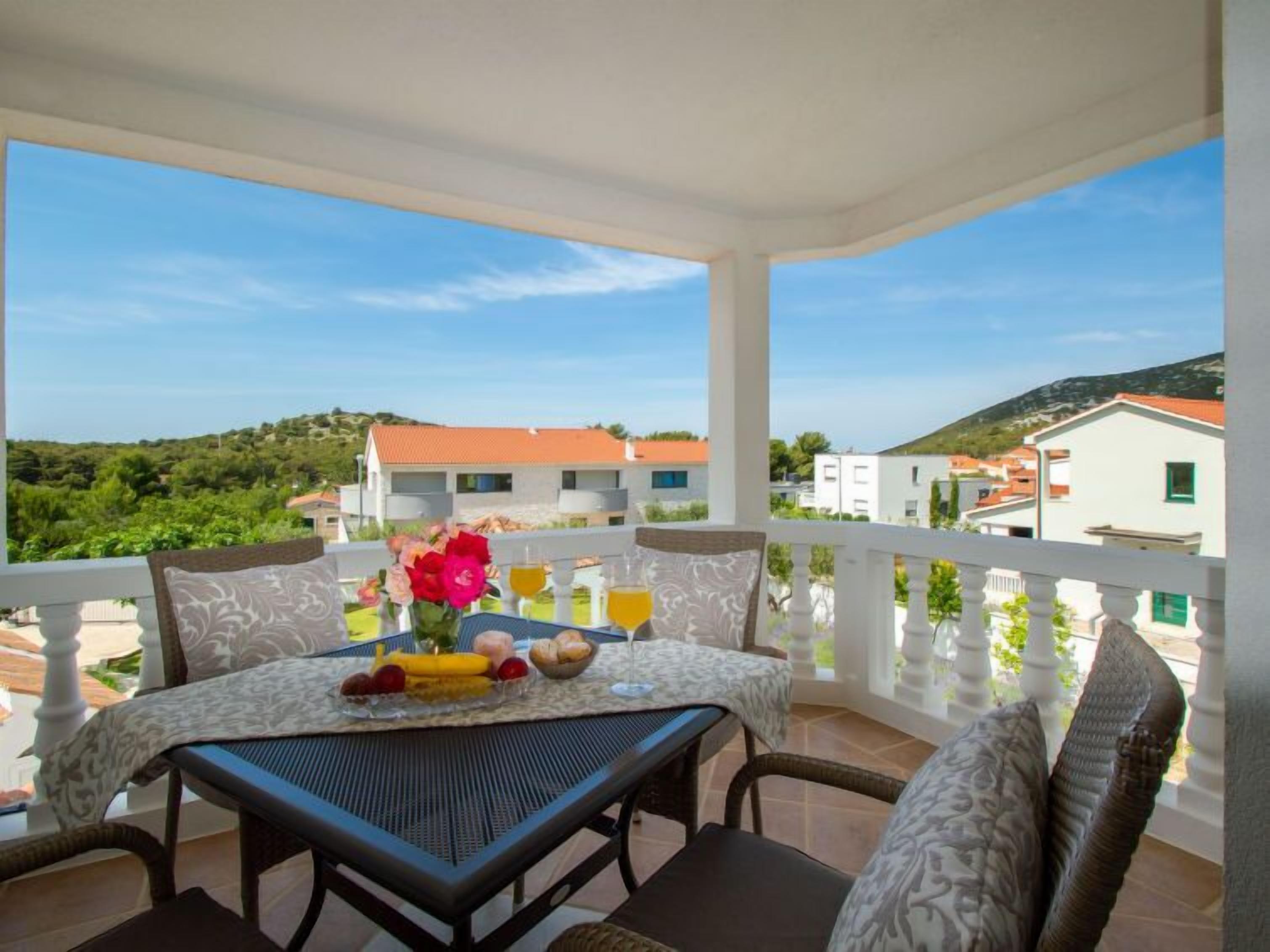 Ein Balkon mit Frühstückstisch, ausgestattet mit Früchten und Getränken, mit Blick auf eine Wohngegend mit Häusern und Grünflächen.