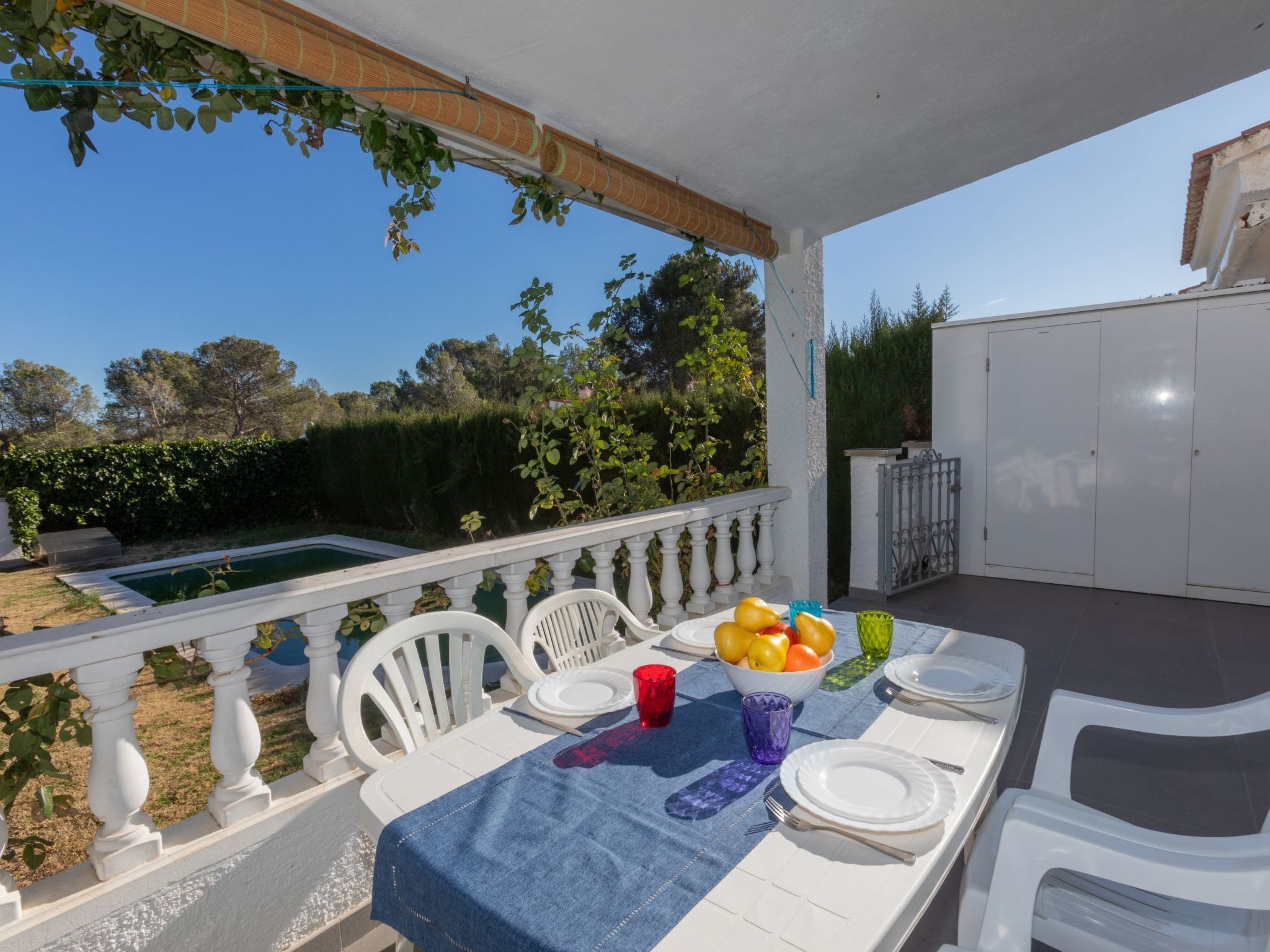 An outdoor dining area with a table set for two, surrounded by white chairs, a pool, and a view of the garden.