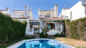 A swimming pool with a clear blue water surface, surrounded by white buildings with red-tiled roofs and a white fence.