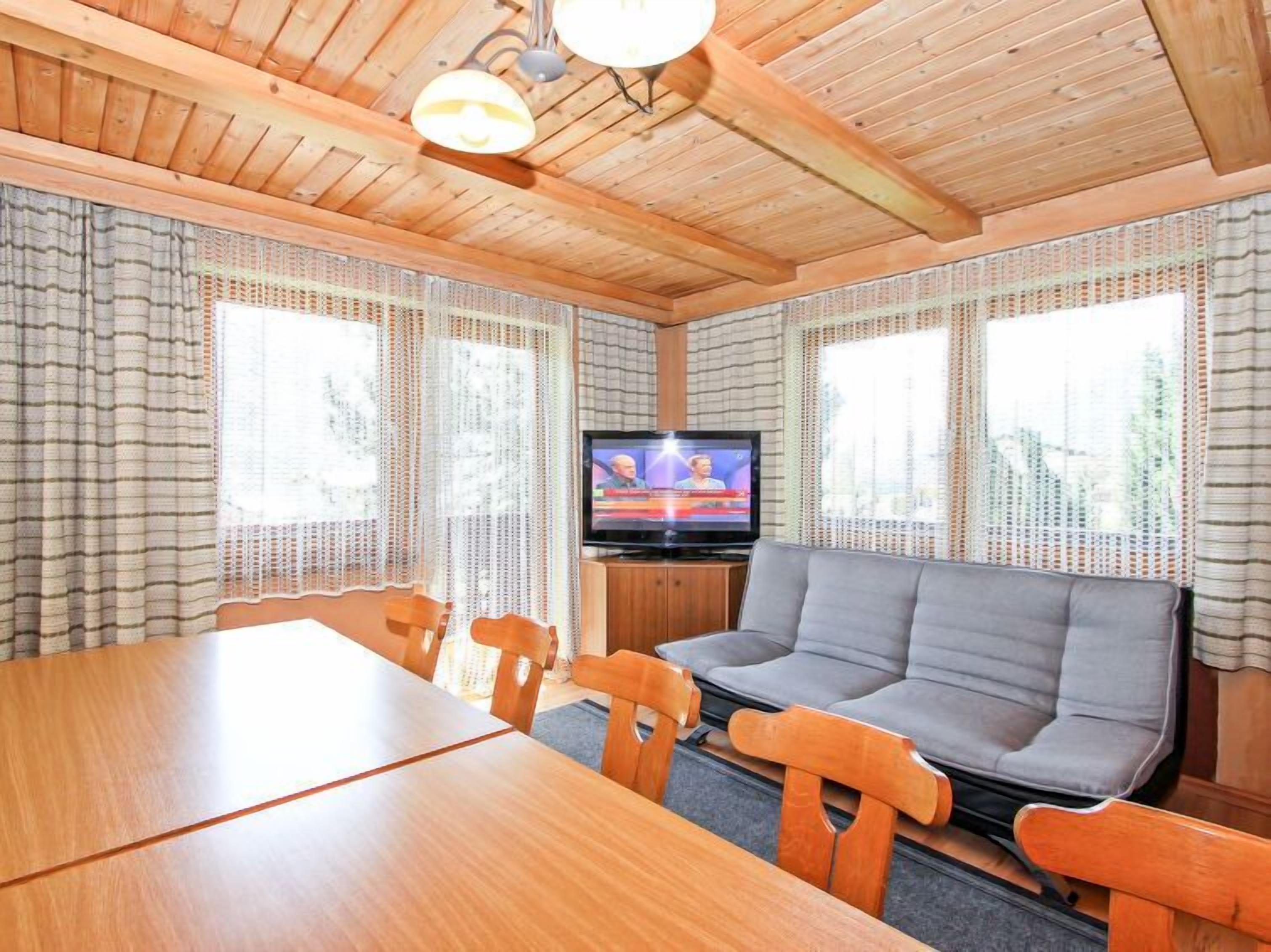 Ein gemütliches Blockhauszimmer mit Holzbalkendecke, einem Fernseher, einer Couch und einem Esstisch mit Stühlen.