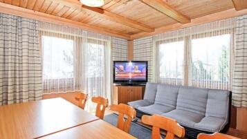 Ein gemütliches Blockhauszimmer mit Holzbalkendecke, einem Fernseher, einer Couch und einem Esstisch mit Stühlen.