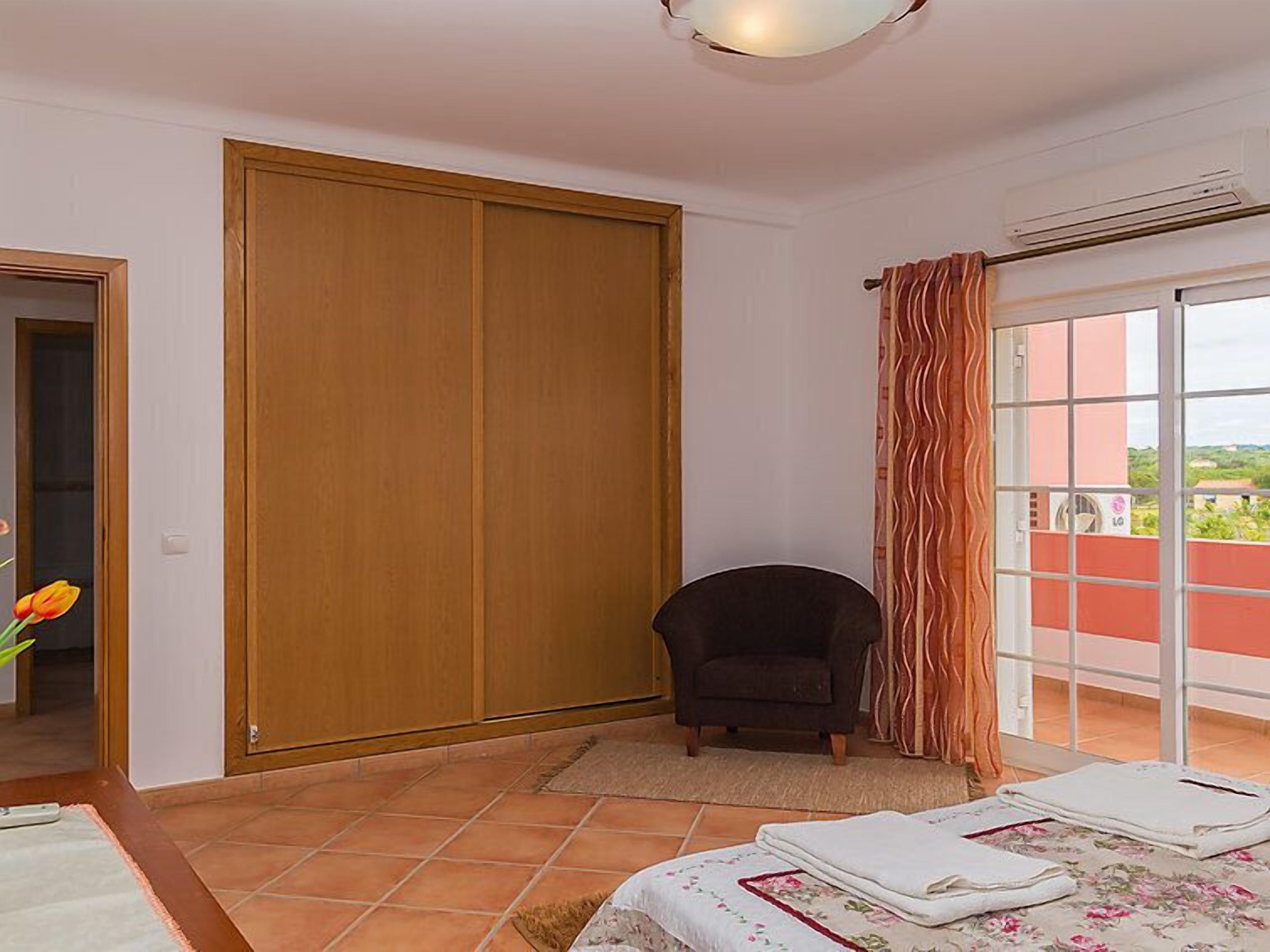 Un dormitorio con un armario grande, una silla, una cama con colcha estampada y vistas a través de una ventana con cortinas.