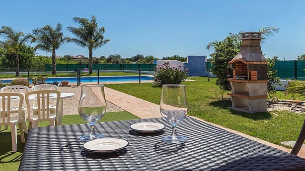 An outdoor dining area with a table set for two, a barbecue grill, and a pool in the background. - Das Amendoeiras by Interhome (Alcantarilha)