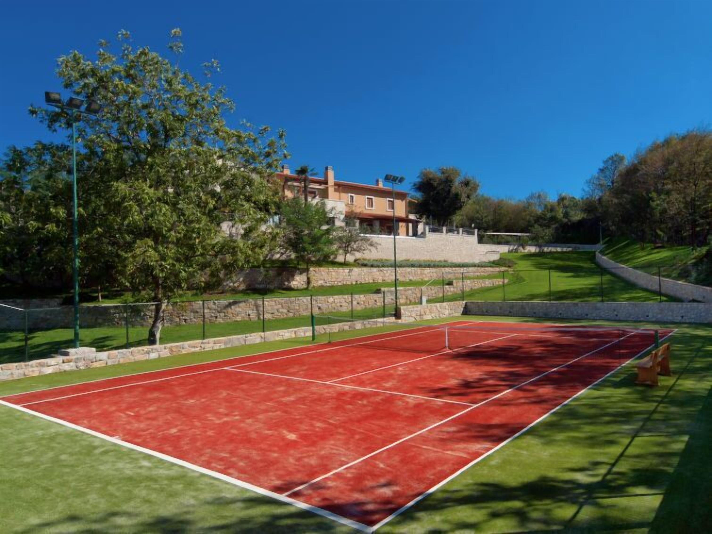 Ein Tennisplatz mit roter Belag, umgeben von einer Steinmauer und Grünflächen, mit einem Gebäude im Hintergrund.