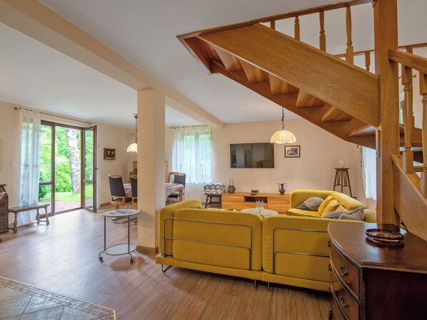 A spacious living area with a wooden staircase, a yellow sofa, a dining table, and a TV mounted on the wall.