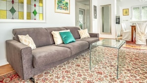 Een woonkamer met een grijze bank, een glazen salontafel en een vloerkleed met bloemenprint.
