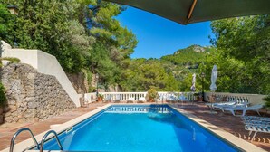 A rectangular swimming pool with a stone wall on one side, surrounded by lounge chairs and umbrellas, set against a backdrop of trees and hills.