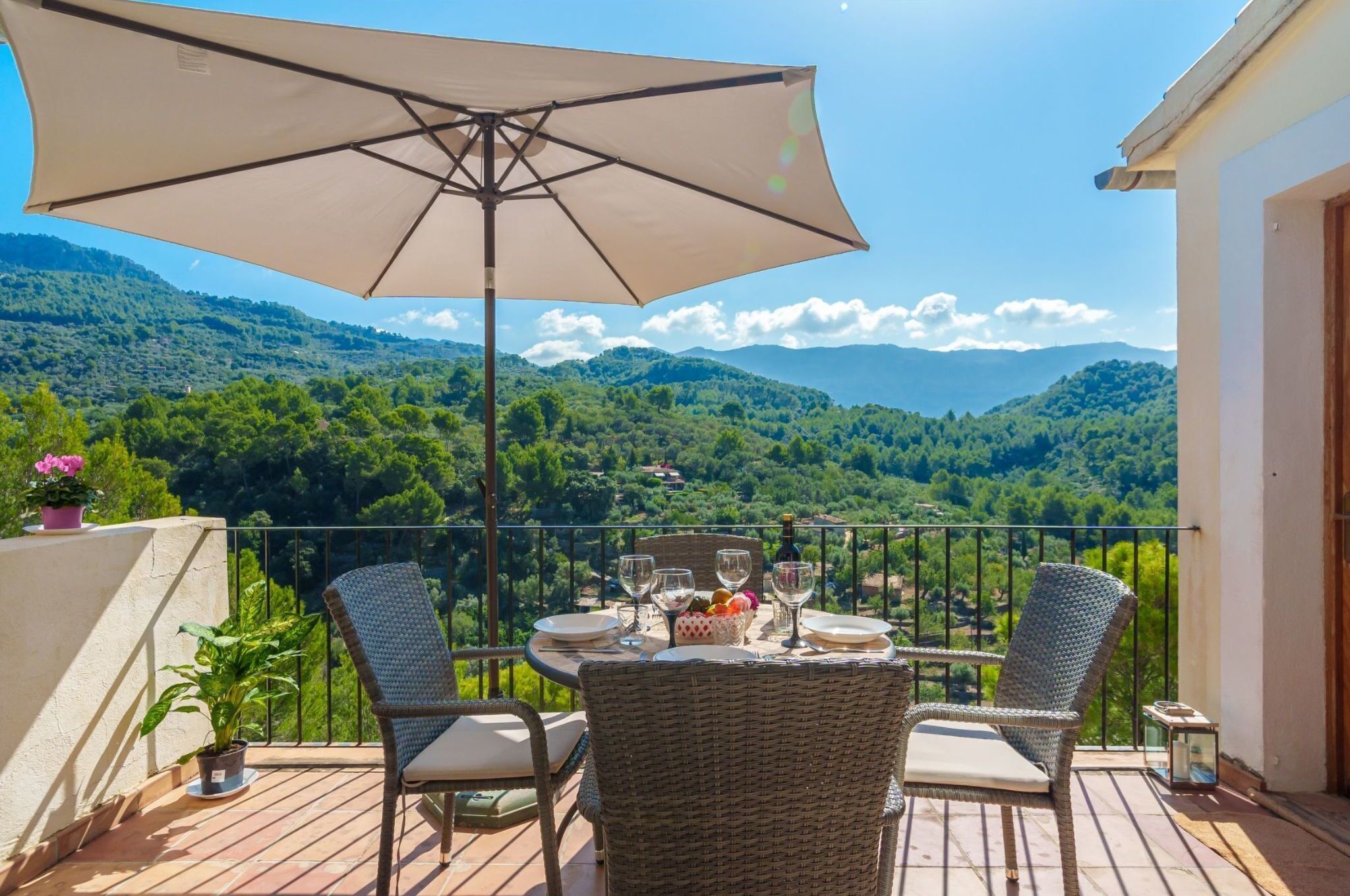 Un balcon avec des fauteuils en osier, une table dressée avec des assiettes, des verres et une bouteille, et un grand parasol.