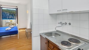 A compact room with a bed, a small table with chairs, a kitchen with a sink and stove, and a view of a coastal landscape through a window. - Apartment Cico by Interhome (Hvar/Pokrivenik)