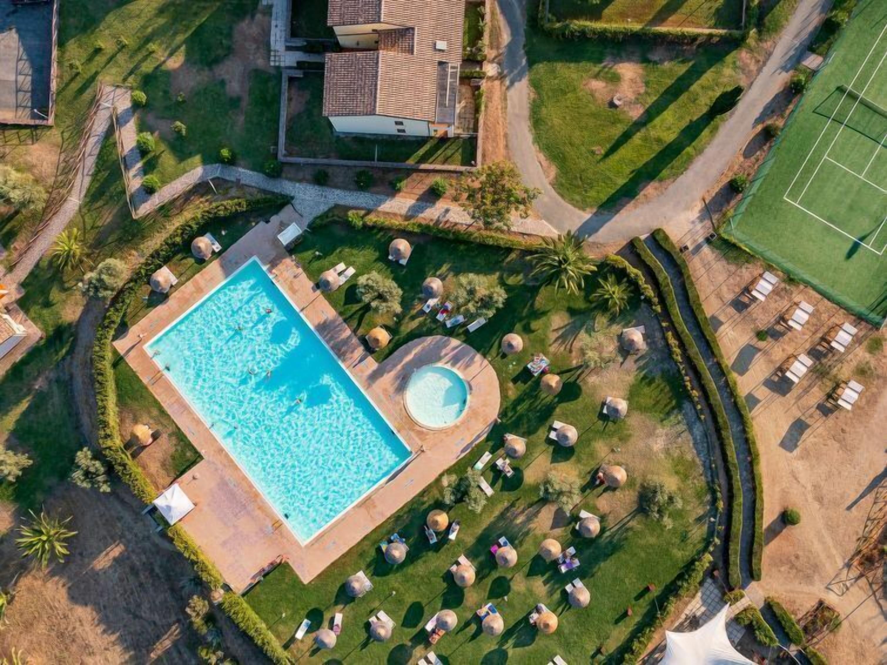 An aerial view of a resort with a large rectangular swimming pool, a circular hot tub, and several outdoor seating areas with umbrellas.