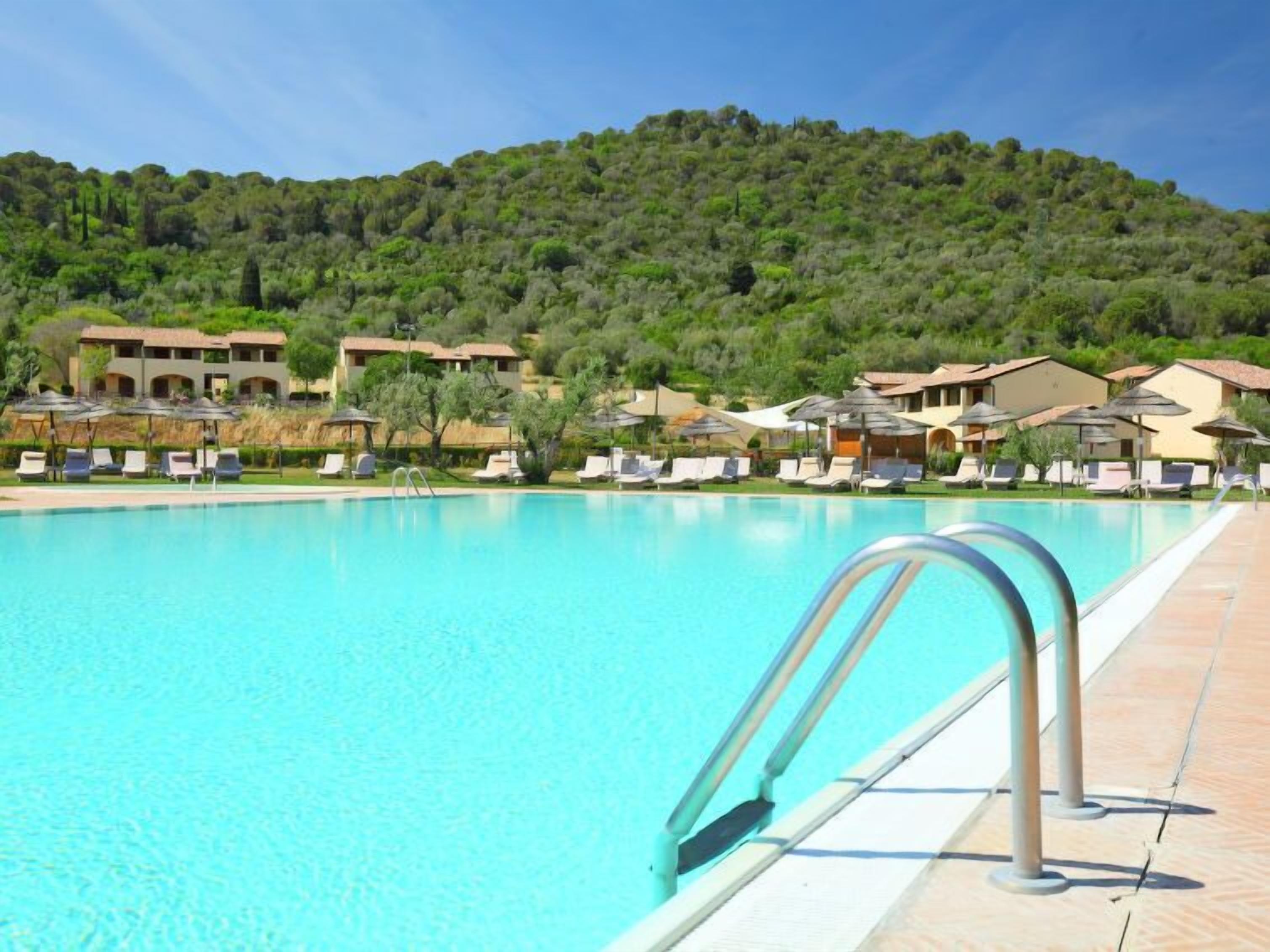 A swimming pool with lounge chairs and umbrellas, surrounded by buildings with a mountainous backdrop.