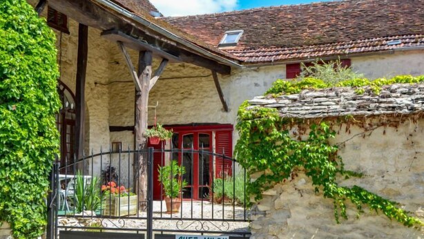A rustic stone building with a red door, a black metal gate, and a sign reading "CHEZ MILOU".