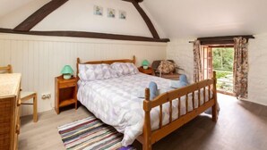 A bedroom with a sloped ceiling, wooden beams, a bed with a checkered bedspread, a nightstand with a lamp, a chair, a dresser, and a window with curtains.