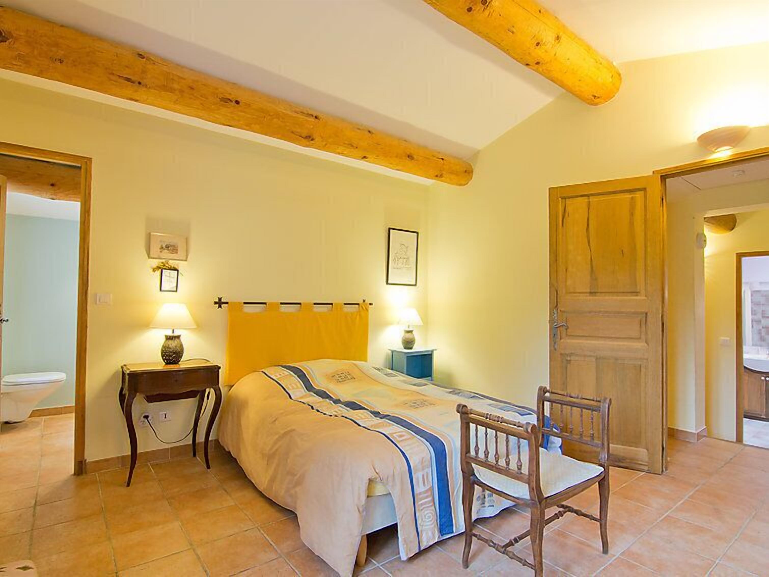 Ein Schlafzimmer mit einem Bett, Nachttischen, einem Sessel, einer Holzbalkendecke und einer Tür.