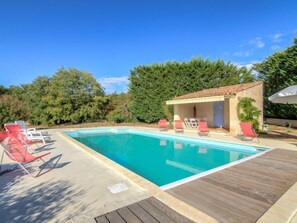 Una piscina rectangular con agua azul cristalina, rodeada por una terraza de madera. Hay varias sillas de descanso rojas y una pequeña zona cubierta con techo de azulejos.