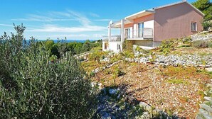 Una casa de dos pisos con un balcón, rodeada de olivos y un paisaje rocoso.