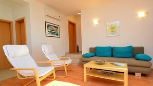 Una sala de estar con dos sillones blancos, una mesa de centro de madera y un sofá gris con cojines color turquesa. Hay dos puertas, una con un cuadro encima, y una escalera que lleva a otra habitación.