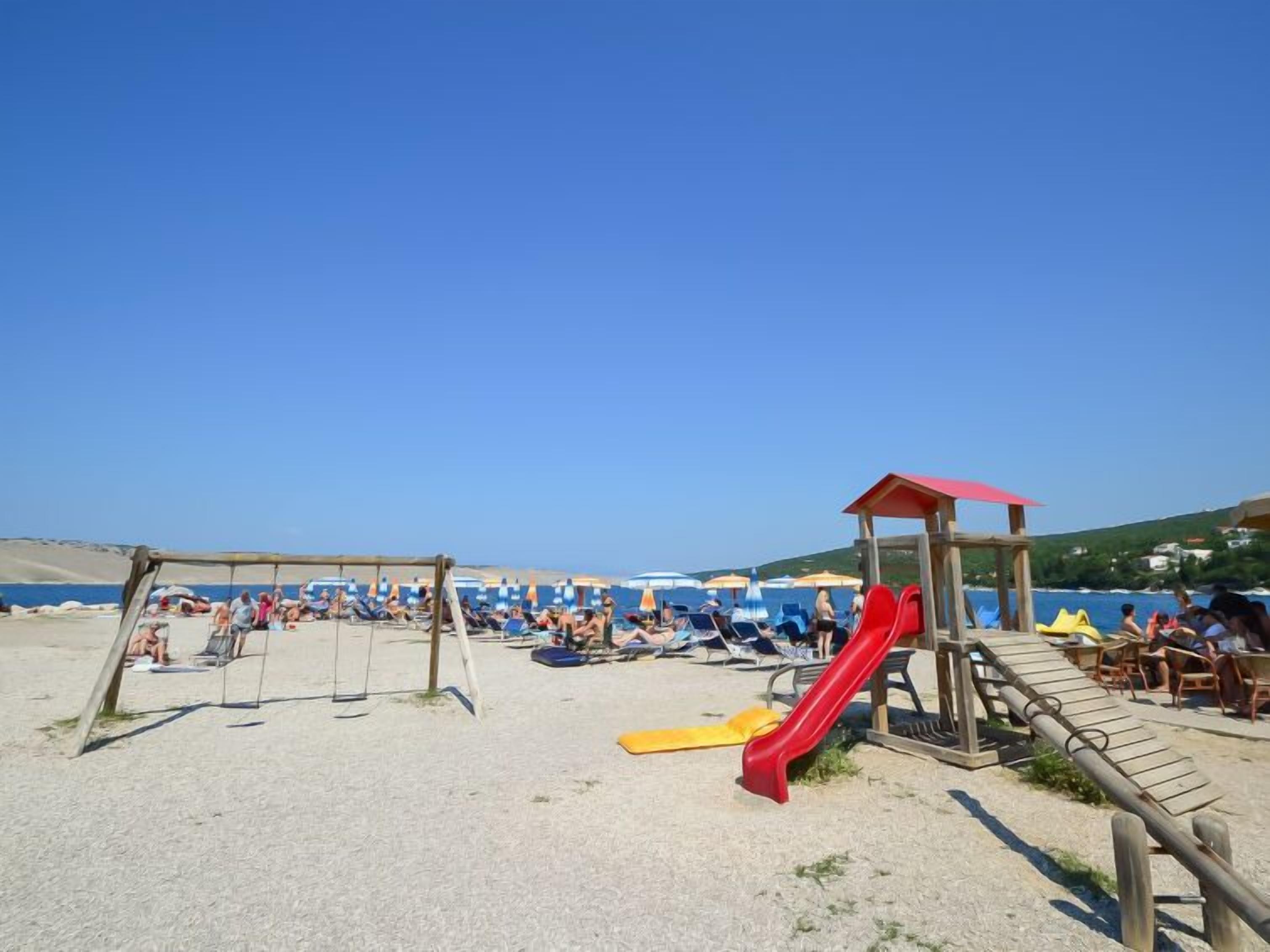 A beach with a red slide, a wooden play structure, and beach chairs under umbrellas.