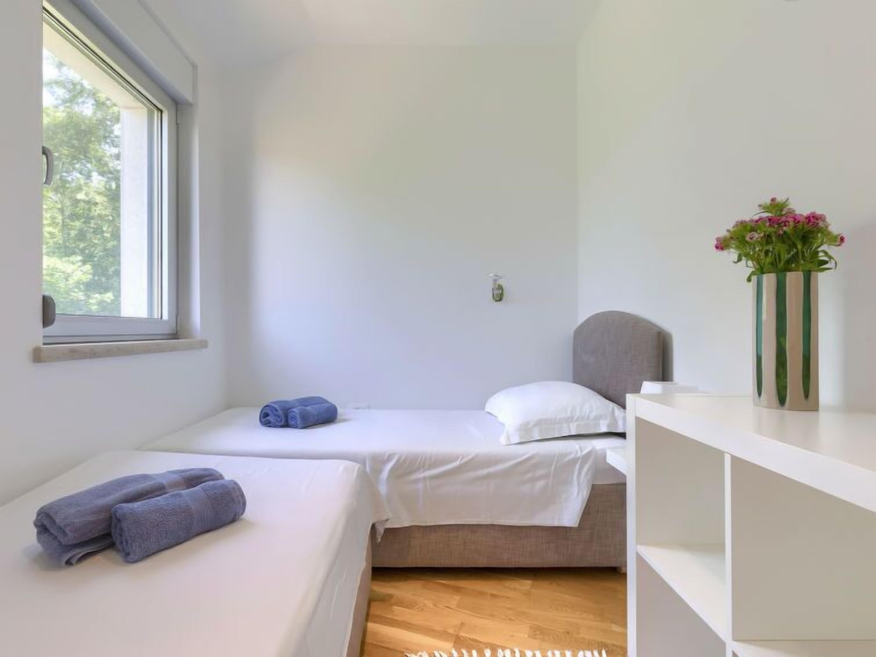 A small, bright room with two beds, a window, and a shelf with a vase of flowers.