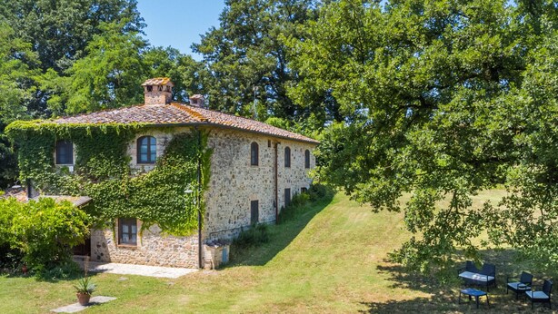 Une maison en pierre avec un toit de tuiles, entourée d’une végétation luxuriante et d’arbres.