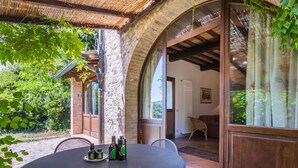 Une table ronde entourée de chaises, avec des bouteilles d’huile d’olive, dans un espace patio extérieur doté de murs en pierre et d’une porte en bois.