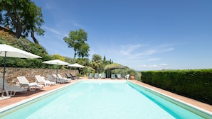 Ein rechteckiger Swimmingpool mit klarem, blauem Wasser, umgeben von weißen Liegestühlen und Sonnenschirmen, vor einer Kulisse aus Bäumen und einer Steinmauer.