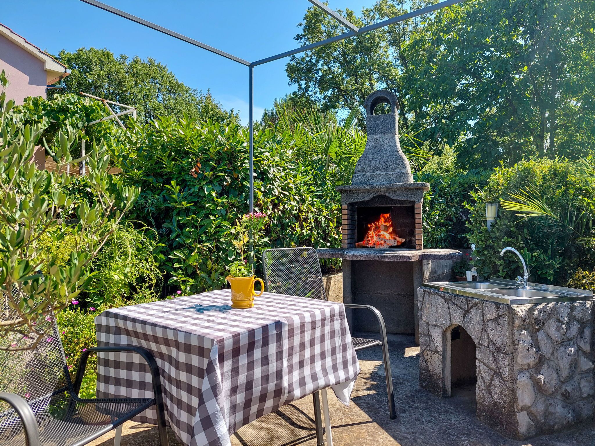 An outdoor patio with a stone-built pizza oven, a table with a checkered tablecloth, and a metal chair.