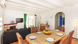 A dining area with a wooden table, chairs, and a fruit bowl. There is a sofa and a television in the background. - Afrodita by Interhome (L'Escala)