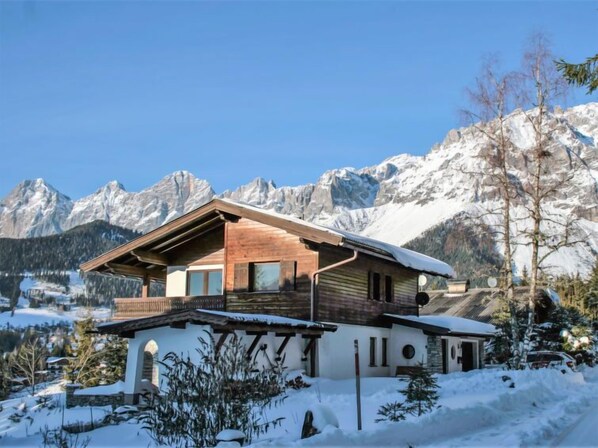 A chalet with a wooden exterior, large windows, and a balcony overlooking a snowy landscape with mountains in the background.