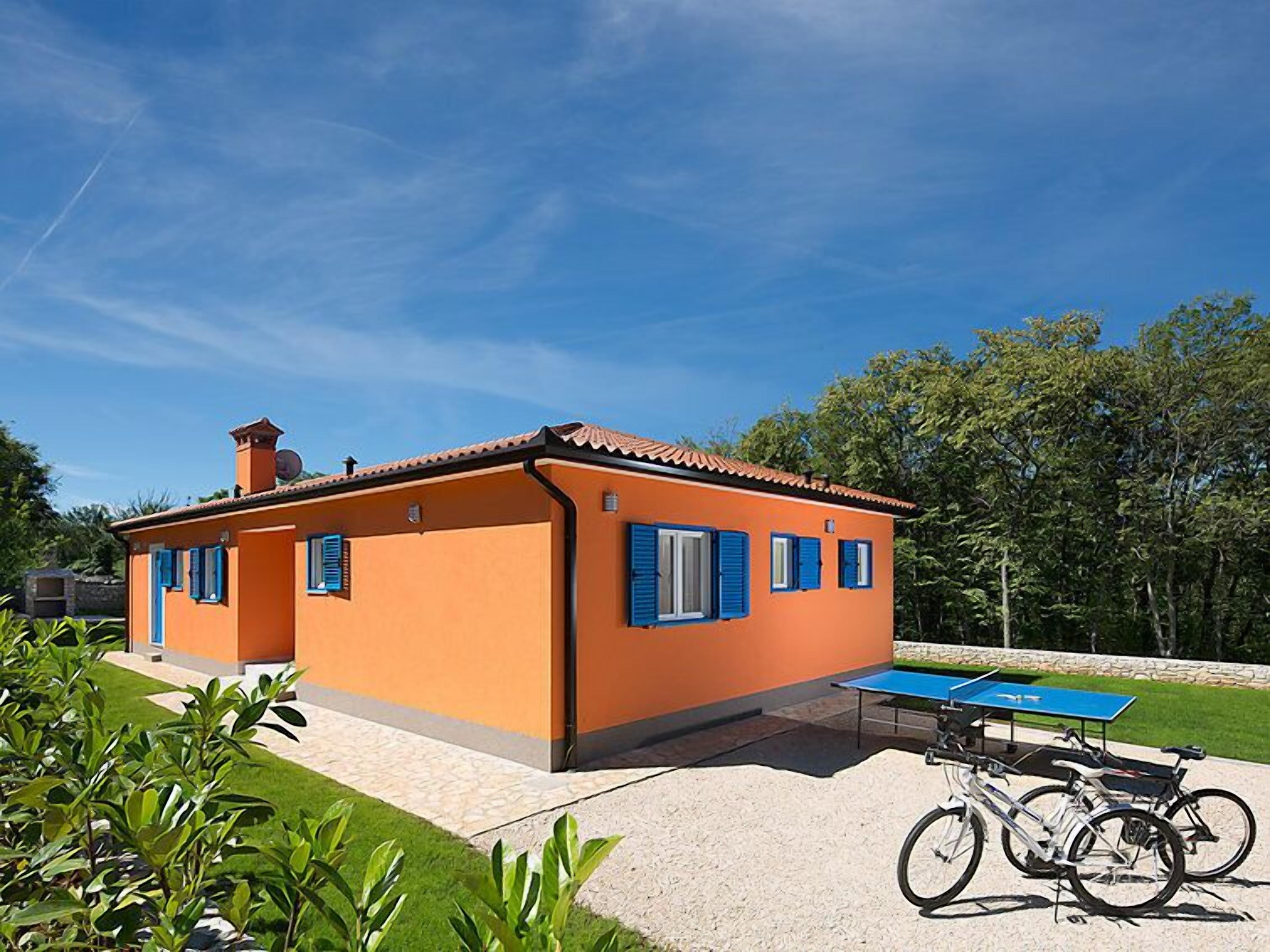 Un bâtiment d’un seul étage, avec un toit en tuiles, des murs orange et des volets bleus. On peut voir une table de ping-pong et des vélos au premier plan.