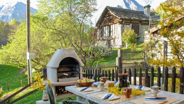 Cozinha externa de pedra com forno a pizza, mesa posta para refeições e vista para uma montanha ao fundo.