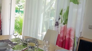 Et spisebord dækket til fire med en moderne pendellampe, hvide gardiner med blomstermønster og udsigt til grønne omgivelser.