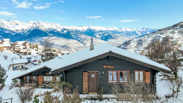 A rustic cabin with a snow-covered roof, wooden exterior, and a chimney. It is surrounded by a snowy landscape with mountains in the background. - Mustela by Interhome (Nendaz)