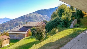 A house with a sloped roof, surrounded by lush greenery and mountains in the background. - Mustela by Interhome (Nendaz)