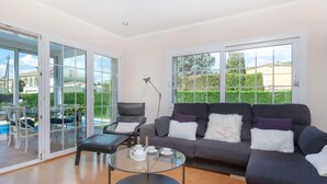 A modern living room with a grey sofa, a glass coffee table, and a chair. There are large windows with a view of a garden and a pool. - Amsterdam by Interhome (St Antoni de Calonge)