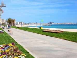 A coastal promenade with a concrete path, grassy areas, and flower beds.