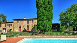 A swimming pool with a stone deck, a stone building with red doors, and a tall green tree in the background.
