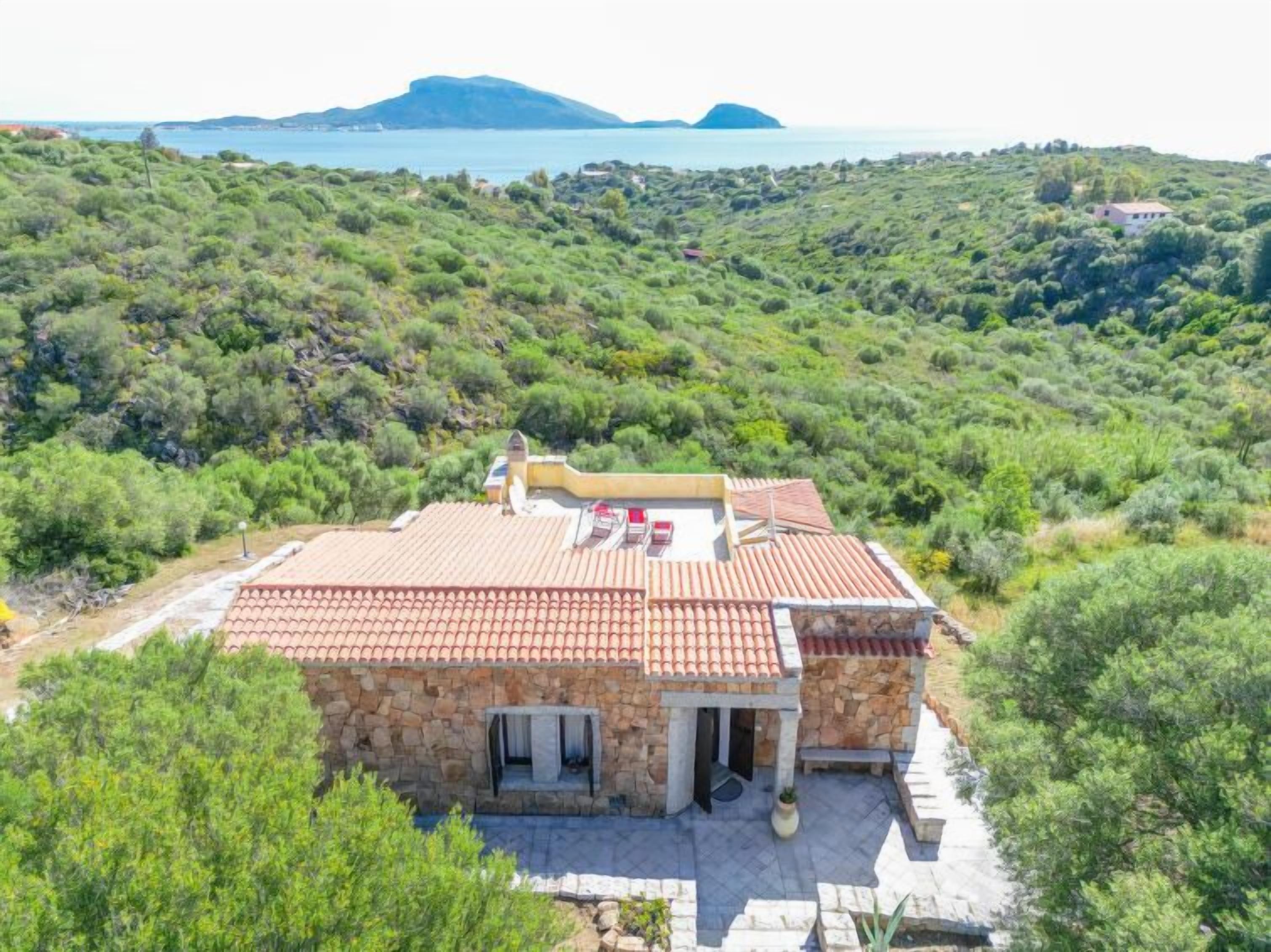 A stone house with a tiled roof, a small porch, and a view of the sea and hills in the background.