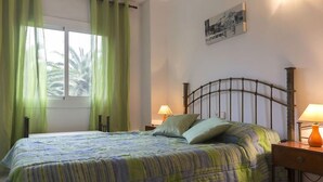 Een netjes opgemaakt bed met een bedsprei met patroon, een nachtkastje met een lamp en een raam met groene gordijnen.