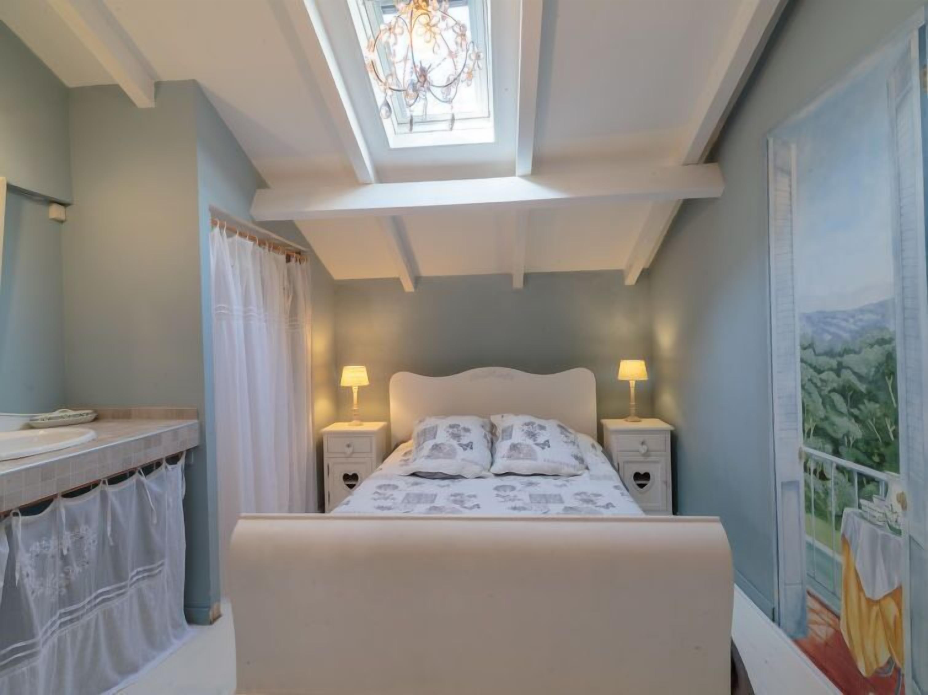 A bedroom with a bed, two bedside tables with lamps, a window with a view, and a ceiling light fixture.
