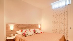 A bedroom with a wooden headboard, two bedside lamps, a bed with floral bedding, a small table, and a patterned curtain. - Apartment Laurus by Interhome (Lovran/Tuliševica)