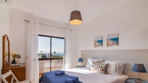 Un dormitorio con una cama grande, vista al mar, un escritorio con lámpara y una silla.