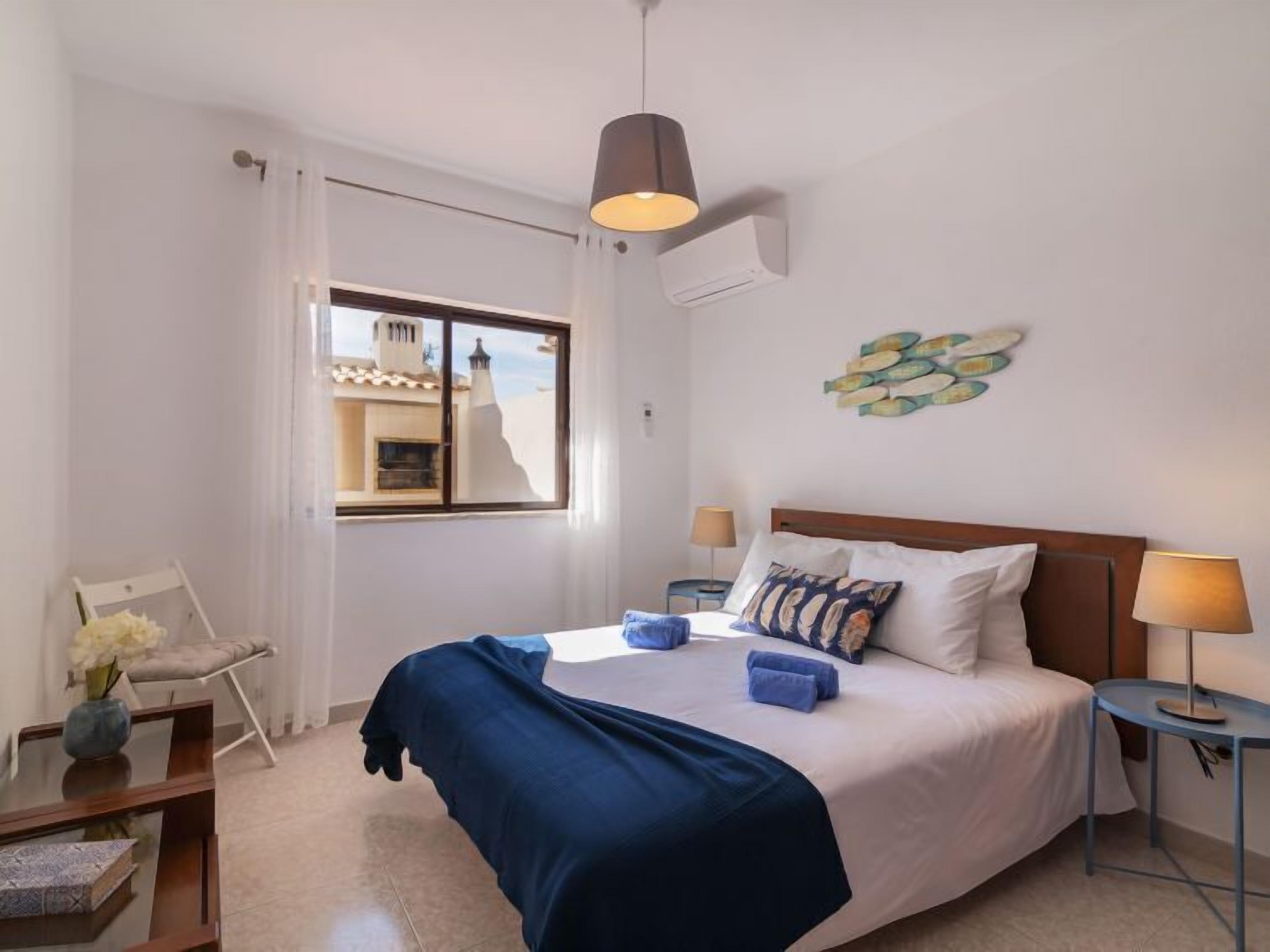 A bedroom with a bed, bedside tables, a window with curtains, a wall-mounted air conditioner, and a decorative fish wall art piece.
