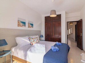 A bedroom with a bed, a bedside table, a lamp, a wardrobe, and a hallway leading to another room.