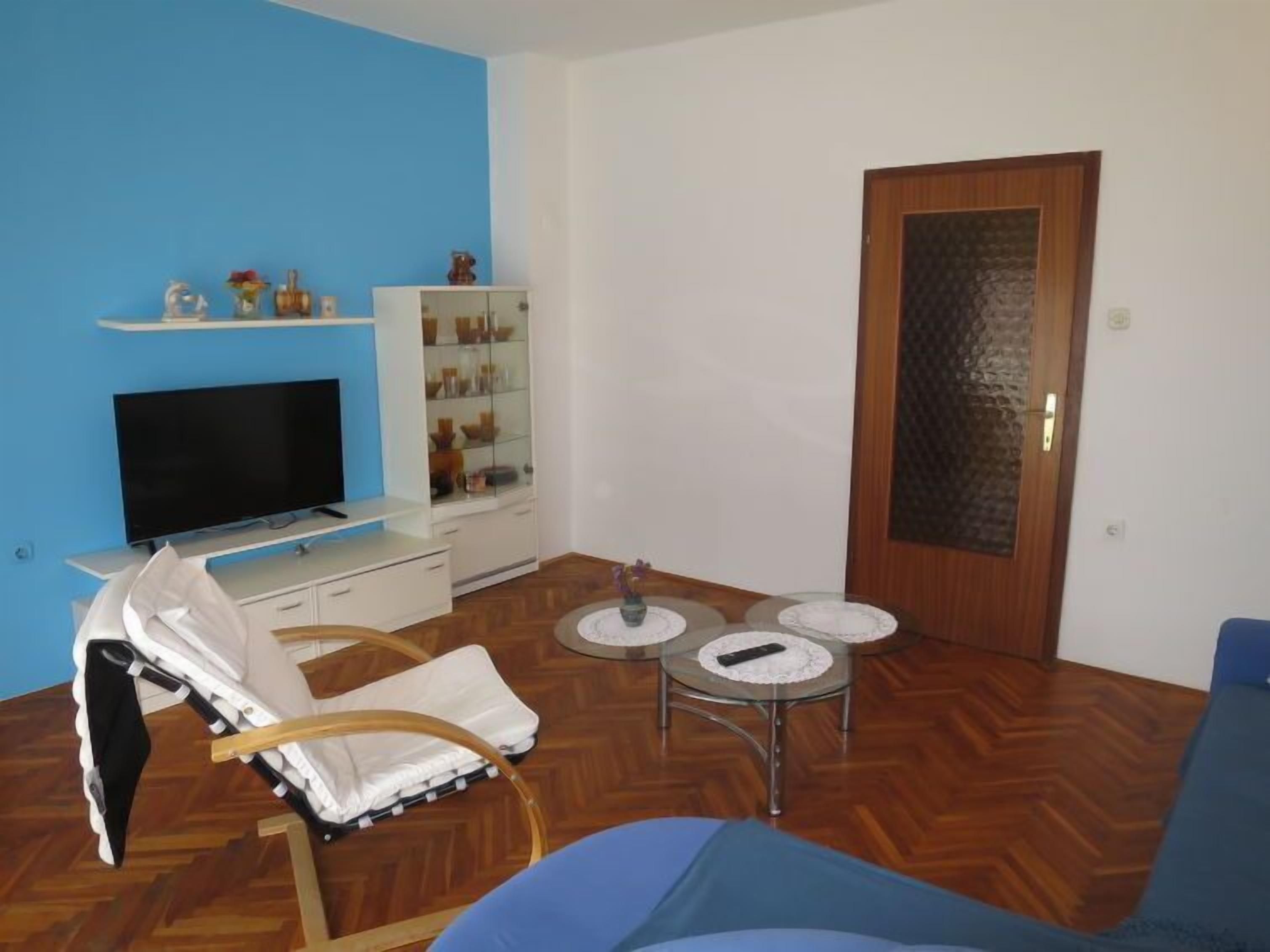 A living room with a flat-screen TV, a white cabinet with glass doors, a glass table with plates, a blue sofa, and a wooden floor.