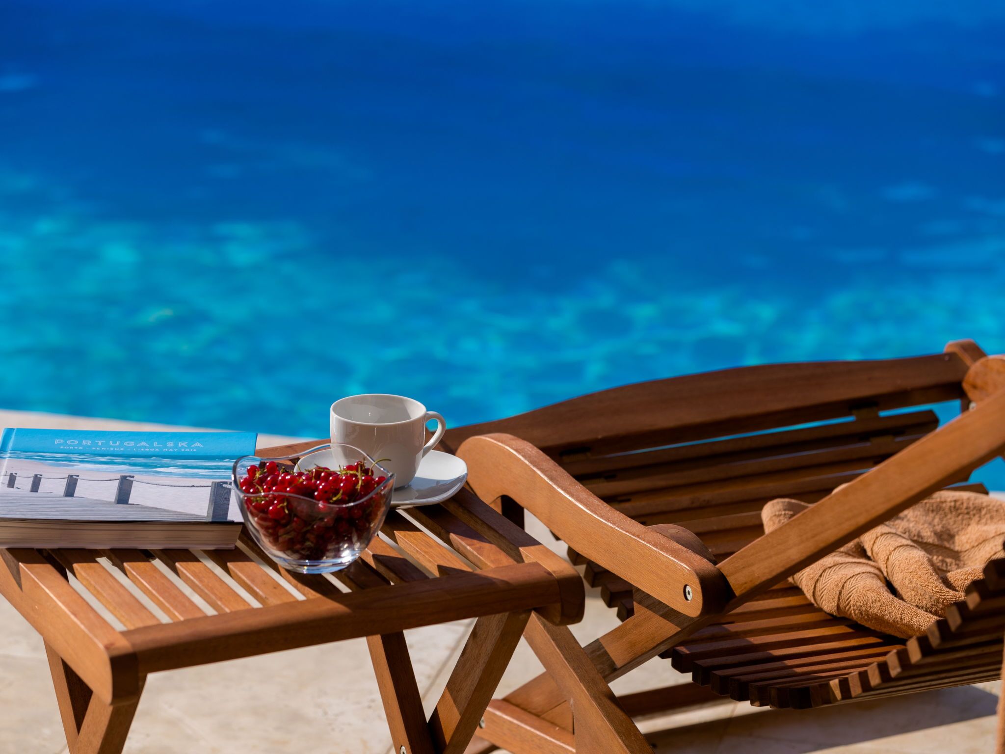 Uma cadeira de lounge de madeira com uma toalha, uma chávena e uma taça de cerejas sobre uma mesa junto a uma piscina.