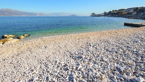 Ein Kiesstrand mit klarem Wasser und Bergen im Hintergrund.
